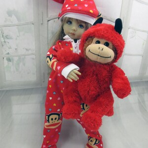 May include: A doll wearing a red onesie and hat with a heart pattern, holding a red monkey plush toy. The onesie features a Paul Frank monkey graphic. The hat has a white trim and a pom-pom.