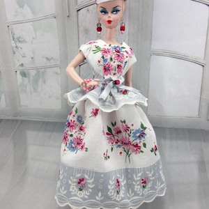 May include: A doll wearing a white dress and matching hat featuring a floral pattern with pink and blue flowers. The dress has a peplum waist with a light blue ribbon tie. The doll wears red and grey drop earrings and a matching bracelet.