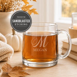 May include: A clear glass mug filled with amber liquid, featuring a sandblasted etching of a large cursive letter M above the name MEGAN. A circular badge overlay on the image displays the text PREMIUM SANDBLASTED ETCHING in white font.