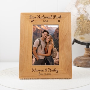 Può includere: Una cornice in legno chiaro con il testo "Zion National Park Utah" e "Warren & Hailey June 12, 2026". La cornice contiene una foto di una coppia. Un mappamondo decorativo è sullo sfondo.
