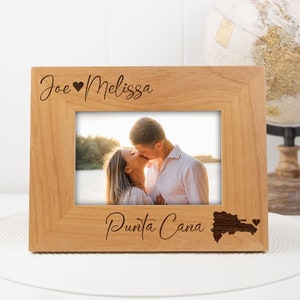 May include: A wooden photo frame with the names "Joe ♥ Melissa" and "Punta Cana" engraved on it. The frame features a photo of a couple kissing in front of a beach and a small outline of the Dominican Republic with a heart.