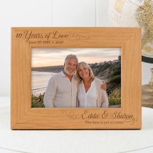 May include: A light-colored wooden picture frame with engraved text reading 40 Years of Love June 27, 1987 - 2027 at the top and Eddie & Sharon The best is yet to come at the bottom, containing a photograph of a couple standing by the ocean.