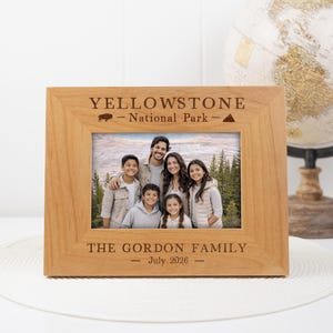 May include: A wooden picture frame with a family photo inside. The frame is engraved with "YELLOWSTONE National Park" and "THE GORDON FAMILY July 2026". A globe is in the background.
