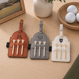 May include: Three rectangular golf bag tags in tan, gray, and white leather, each featuring a buckle strap, a horizontal pocket holding three wooden golf tees, and custom black monogrammed logos including initials and a golf club graphic.