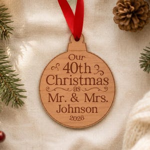 Op de afbeelding: Een ronde houten kerstornament met een rood lint. Het ornament is gegraveerd met de tekst: Our 40th Christmas as Mr. & Mrs. Johnson 2026. Het ontwerp bevat decoratieve krullen boven en onder de centrale tekstregels.