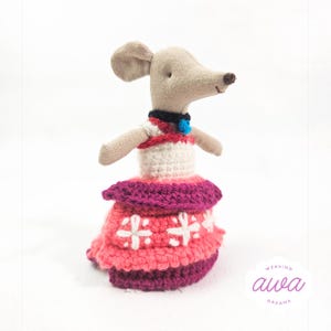 May include: A small crocheted mouse doll wearing a pink and white dress with a blue collar and a blue bell. The doll is standing on a white surface. The text "weaving awa dreams" is visible in the lower right corner of the image.