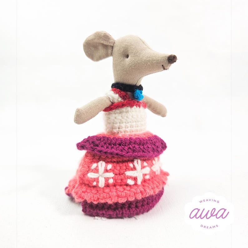 May include: A small crocheted mouse doll wearing a pink and white dress with a blue collar and a blue bell. The doll is standing on a white surface. The text "weaving awa dreams" is visible in the lower right corner of the image.