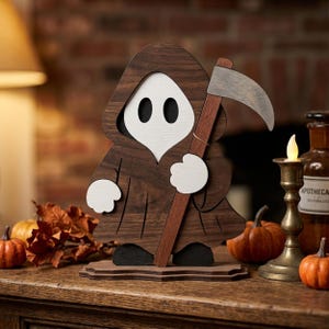 May include: A wooden figure of a hooded character with a white face and black eyes, wearing a brown robe and holding a gray scythe. The figure stands on a flat base and features white hands, dark feet, and a layered construction with visible wood grain.