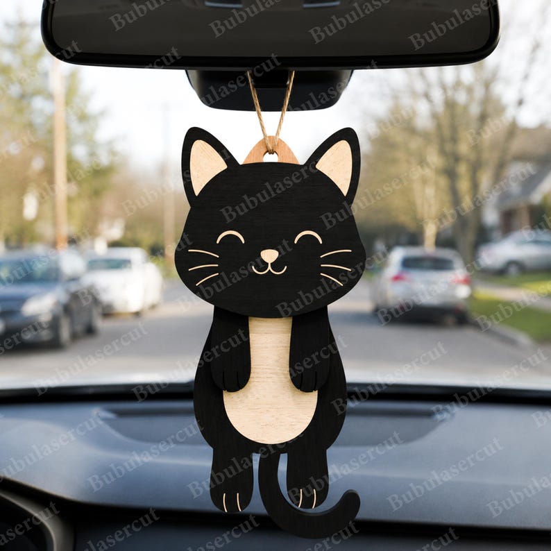 May include: A black cat-shaped car charm with a smiling face and a light-colored belly. The wooden charm hangs from a string and is designed to be suspended from a car's rearview mirror. The charm has a simple, clean design.