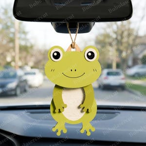 May include: A green frog-shaped car air freshener hanging from a rearview mirror. The frog has large, round eyes, a smiling mouth, and small arms and legs. The air freshener is made of wood and has a light-colored center.