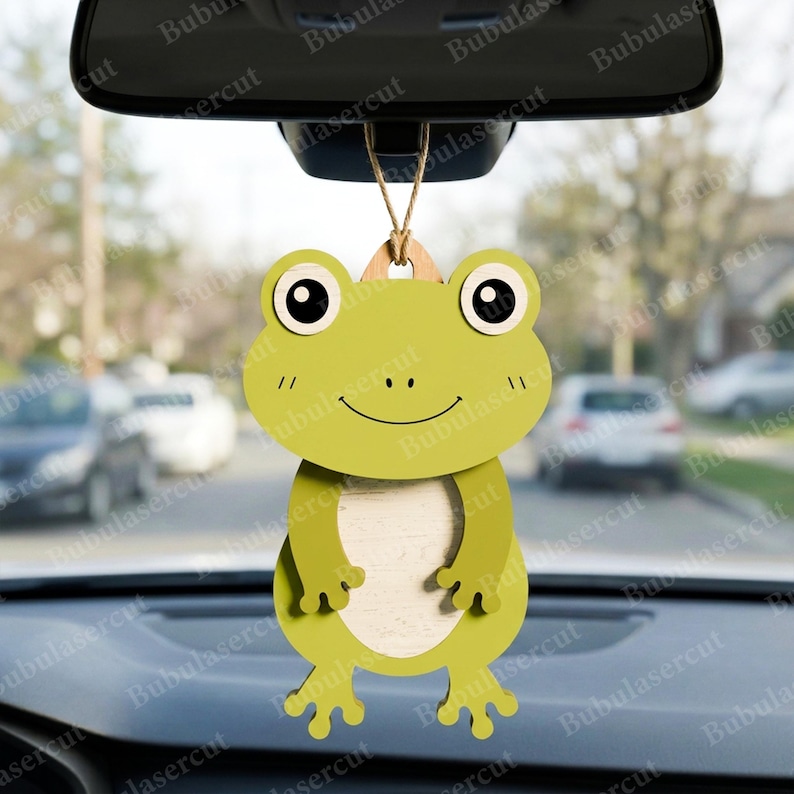 May include: A green frog-shaped car air freshener hanging from a rearview mirror. The frog has large, round eyes, a smiling mouth, and small arms and legs. The air freshener is made of wood and has a light-colored center.