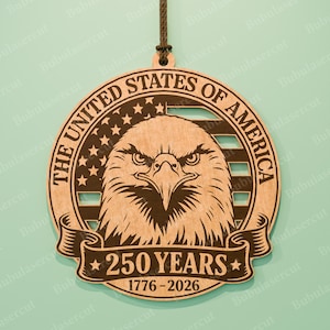 K&ouml;nnte beinhalten: Holzornament mit einem Adler-Design, dem Text "THE UNITED STATES OF AMERICA" und "250 YEARS 1776-2026". Das Design beinhaltet Sterne und Streifen. Das Ornament ist aus hellbraunem Holz gefertigt und hat eine braune Schnur zum Aufh&auml;ngen.