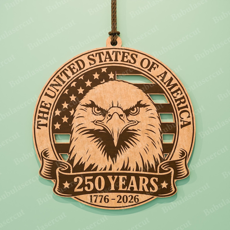 K&ouml;nnte beinhalten: Holzornament mit einem Adler-Design, dem Text "THE UNITED STATES OF AMERICA" und "250 YEARS 1776-2026". Das Design beinhaltet Sterne und Streifen. Das Ornament ist aus hellbraunem Holz gefertigt und hat eine braune Schnur zum Aufh&auml;ngen.