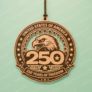 K&ouml;nnte beinhalten: Runder Holzschmuck mit der Aufschrift "UNITED STATES OF AMERICA" und "250 YEARS OF FREEDOM". Zeigt einen Adler, Sterne und die Jahre 1776-2026. Enth&auml;lt eine braune Schnur zum Aufh&auml;ngen. Eine patriotische Dekoration.