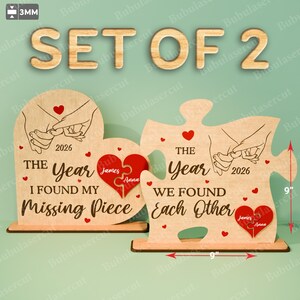以下が含まれることがあります： 「SET OF 2」の文字が入った、ベース付きの木製パズルピース2個セット。片方は「I FOUND MY MISSING PIECE」、もう片方は「WE FOUND EACH OTHER」と書かれています。各ピースには赤いハートと、名前入りのパズルピース型のハートが付いています。