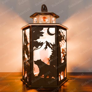 May include: A wooden lantern with a cut-out design of a bear, mountains, and trees. The lantern is lit from inside, casting a warm glow.