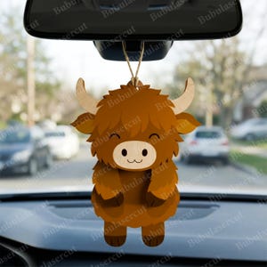 May include: A wooden car charm featuring a cartoon Highland cow design. The charm is brown with a light wood face and horns, and is suspended by a natural twine. The background shows a car interior and a street scene.