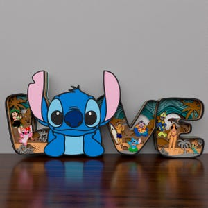 May include: A wooden cutout of the word 'LOVE' with a blue Stitch character in the center of the 'O'. The letters are decorated with a beach theme, including palm trees, waves, and sand. There are also small characters from the Lilo & Stitch movie, including Stitch, Jumba, and Pleakley.