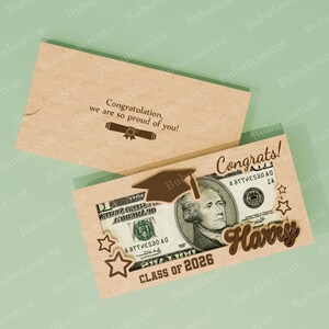 Pu&ograve; includere: Biglietto di laurea in legno marrone chiaro con una banconota da un dollaro visibile. La carta presenta le parole "Congrats!" e "Harry", insieme a un cappello di laurea e stelle. &Egrave; presente anche il testo "Class of 2026".