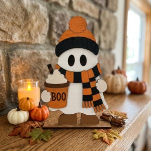 May include: A white wooden ghost cutout decoration wearing an orange and black beanie and a matching striped scarf. The ghost holds a brown cup with a white lid and the text BOO printed on the front. The figure stands on a flat wooden base.