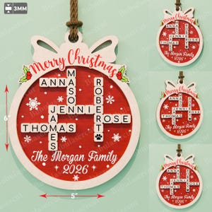 May include: A round wooden Christmas ornament measuring 15.24 cm by 12.7 cm and 3mm thick. The red centre features a crossword-style grid of names, the text Merry Christmas, The Morgan Family, and 2026, decorated with white snowflakes and holly.