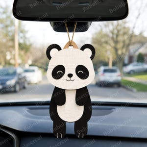 May include: A wooden panda bear car charm with a smiling face, black ears, and black and white body. The charm is hanging from a string, designed to be hung from a car's rearview mirror. The background shows a street scene.