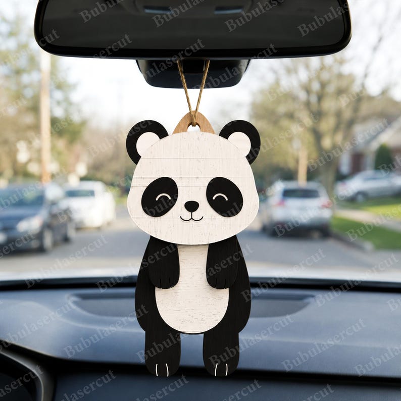 May include: A wooden panda bear car charm with a smiling face, black ears, and black and white body. The charm is hanging from a string, designed to be hung from a car's rearview mirror. The background shows a street scene.