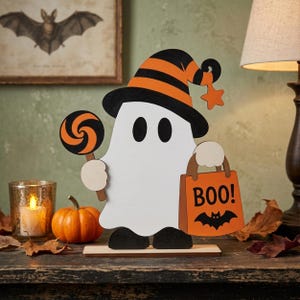 May include: A white ghost cutout decoration wearing a black and orange striped hat, holding an orange and black swirl lollipop in one hand and an orange trick-or-treat bag with the text BOO! and a black bat graphic in the other hand.