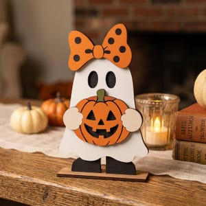 May include: A freestanding wooden ghost decoration featuring a white body, black feet, and an orange bow with black polka dots. The ghost holds an orange jack-o'-lantern with a green stem and black cutouts for eyes and a smiling mouth.