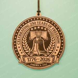 K&ouml;nnte beinhalten: Ein runder Holzschmuck, der 250 Jahre Freiheit feiert. Das Design zeigt die Freiheitsglocke, Lorbeerbl&auml;tter, Sterne und den Text "CELEBRATING 250 YEARS OF FREEDOM" und "1776-2026 AMERICA 250". Ein brauner Faden ist zum Aufh&auml;ngen befestigt.