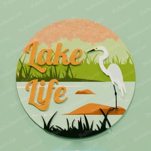 May include: A round wooden sign with a lake scene. The sign features the words "Lake Life" in orange, a white heron, and a lake with grass and rocks. The background is green and light blue.