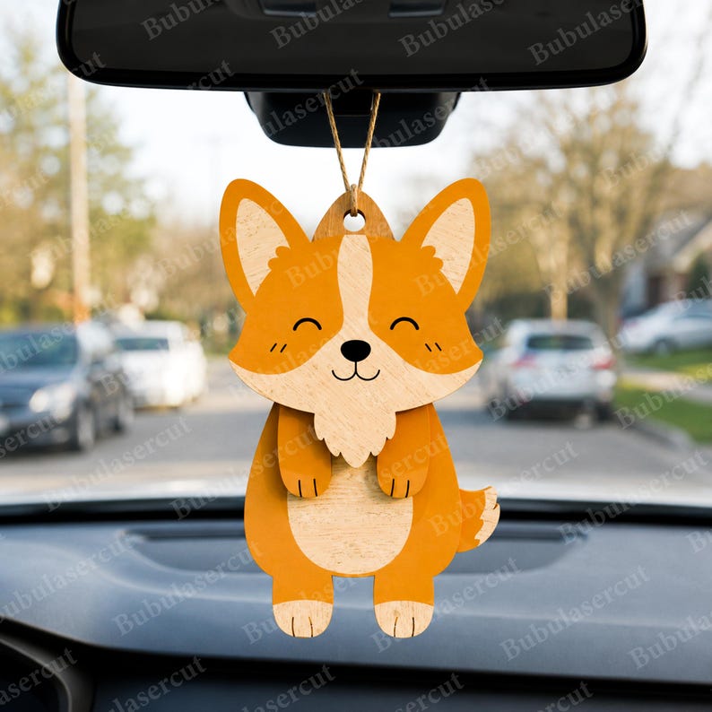 May include: A wooden corgi car charm with orange and natural wood tones. The charm features a smiling corgi face and body, with a small loop for hanging. The charm is suspended from a rearview mirror.