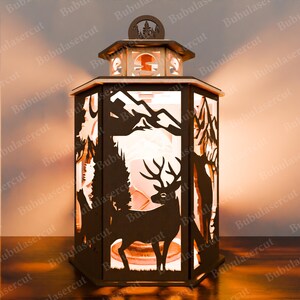 May include: A wooden lantern featuring a cut-out design of a deer standing in a forest scene. The lantern is lit from within, casting a warm glow.