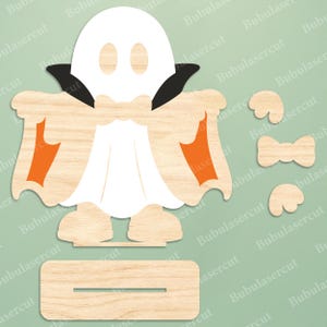 May include: A laser-cut wooden ghost figure wearing a black cape with orange accents, featuring a white ghost head, a separate wooden base with a slot, and additional small wooden bow tie and wing-shaped pieces for assembly and decoration.