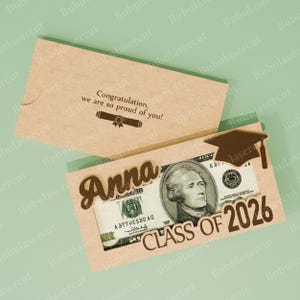 Pu&ograve; includere: Un biglietto di laurea in legno con il nome "Anna" e "CLASS OF 2026" ritagliati. Una banconota da dieci dollari &egrave; visibile attraverso i ritagli. La carta include un disegno di cappello di laurea e il messaggio "Congratulation, we are so proud of you!"