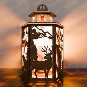 May include: A wooden lantern featuring a cut-out design of a deer standing in a forest scene. The lantern is lit from within, casting a warm glow.