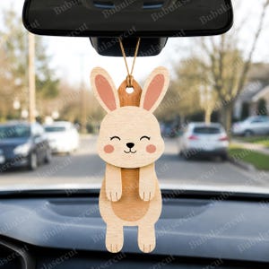 May include: Wooden bunny car charm with a smiling face, pink cheeks, and pink-lined ears. The bunny is hanging from a string, designed to be hung from a car's rearview mirror. The charm is made of light and dark wood.