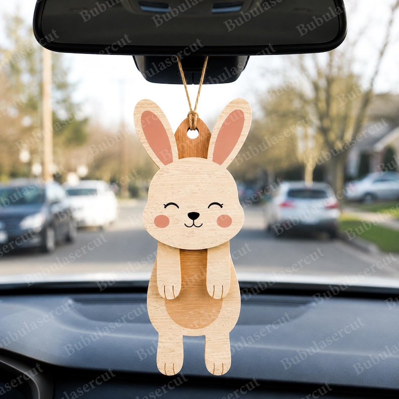 May include: Wooden bunny car charm with a smiling face, pink cheeks, and pink-lined ears. The bunny is hanging from a string, designed to be hung from a car's rearview mirror. The charm is made of light and dark wood.