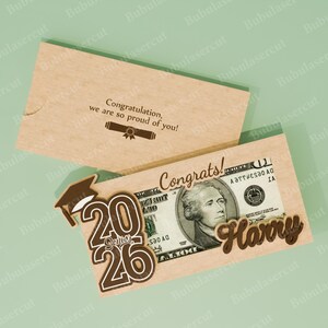 Pu&ograve; includere: Un portadenaro a tema laurea con un tocco marrone, l'anno "2026" e il nome "Harry". Il supporto &egrave; realizzato in cartoncino marrone chiaro e include una banconota da un dollaro. Il testo "Congrats!" &egrave; visibile.
