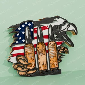 Pu&ograve; includere: Un ritaglio di legno di una bandiera americana con tre proiettili davanti. La bandiera &egrave; strappata e consumata, e i proiettili sono stilizzati con un'aquila e un soldato sullo sfondo. L'aquila &egrave; appollaiata in cima ai proiettili.