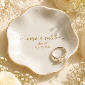 May include: A white, scalloped-edge ceramic ring dish featuring gold-colored text that reads nora & caleb, Married, Apr. 20, 2026. A silver-toned ring with a central stone rests on the surface of the dish, which has a subtle crackle finish.