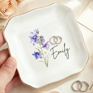 Könnte beinhalten: Eine quadratische weiße Keramikschale mit gewelltem Rand und goldfarbenem Abschluss. In der Mitte befindet sich eine Aquarellillustration von violetten Glockenblumen und der Name Emily in schwarzer Schrift. Zwei Silberringe liegen auf.