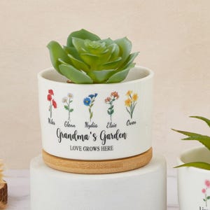 May include: White ceramic planter with a bamboo base. The planter has a floral design with five different flowers and names below each flower. The text reads "Grandma's Garden Love Grows Here".