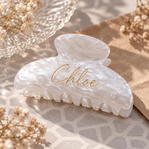 May include: A white hair claw clip with a marbled pattern and the name Chloe written in gold cursive script on the front. The accessory features a curved top edge and a row of interlocking teeth along the bottom for securing hair in place.