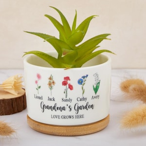 Puede incluir: Maceta de cerámica blanca con base de bambú. La maceta tiene un diseño floral con cinco flores diferentes y los nombres Lionel, Jack, Sandy, Cathy y Avery. El texto "Grandma's Garden" y "Love Grows Here" está impreso en la maceta.