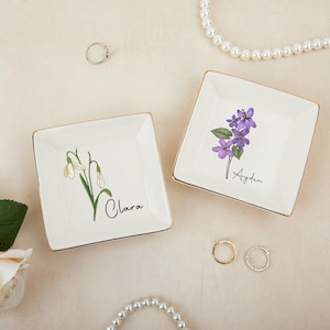 Könnte beinhalten: Zwei quadratische, weiße Keramik-Schmuckschalen mit goldenen Rändern. Eine zeigt eine Schneeglöckchen-Illustration und den Namen "Clara", die andere eine lila Blumenillustration und den Namen "Ayden". Eine Perlenkette und Ringe sind ebenfalls zu sehen.