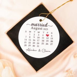 Puede incluir: Adorno de boda redondo blanco con un calendario de agosto de 2025, marcado con un corazón el día 5. El adorno está en una caja negra con una cuerda dorada. El texto "casado/a" y nombres también son visibles.
