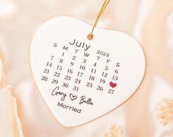 May include: A white heart-shaped ceramic ornament with a gold string for hanging. The ornament features a calendar for July 2025, with the 20th date highlighted in red. The names "Gary & Bella" and the word "Married" are also printed on the ornament.