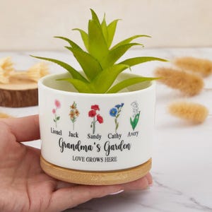 Puede incluir: Maceta de cerámica blanca con base de madera. La maceta tiene un diseño floral con cinco flores diferentes y los nombres Lionel, Jack, Sandy, Cathy y Avery. El texto "Grandma's Garden" y "Love Grows Here" está impreso en la maceta.