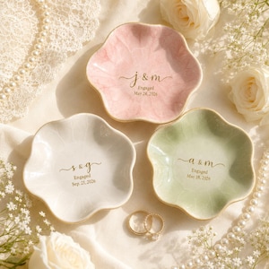 May include: Three flower-shaped ceramic trinket dishes in pastel pink, white, and sage green. Each dish features gold-colored text with initials and engagement dates: j & m May 24, 2026, s & g Sep. 21, 2026, and a & m May 18, 2026. Two gold rings are included.